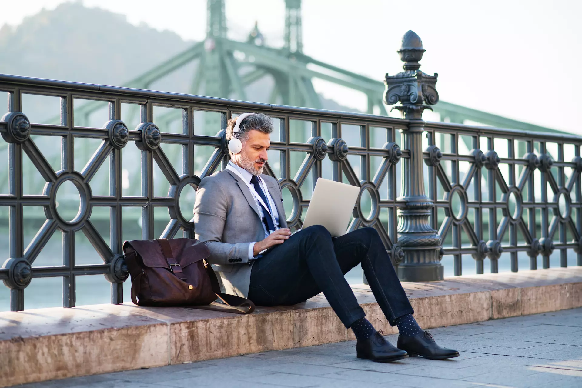 Hübscher reifer Geschäftsmann mit Laptop in einer Stadt. Mann sitzt auf einer Brücke, mit Laptop und Kopfhörern.