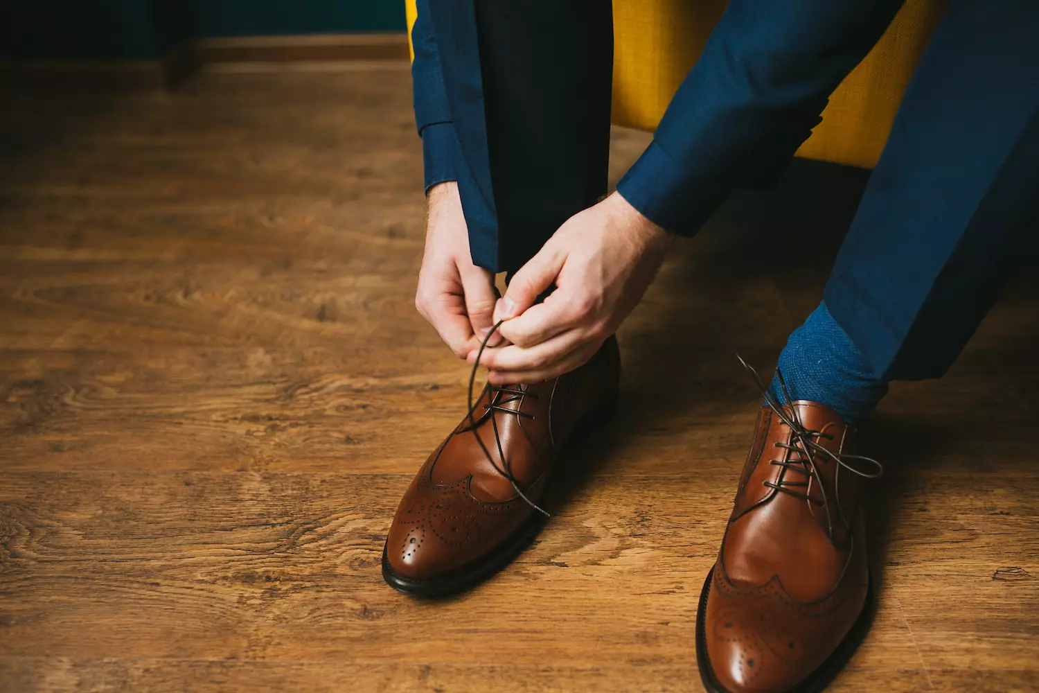 Reconstruíndo o teu garda-roupa: 3 elementos básicos que todo home debería posuír Un home cun traxe azul amarra os cordones dos zapatos nuns zapatos de coiro marróns zapatos brogue sobre un fondo de parquet de madeira