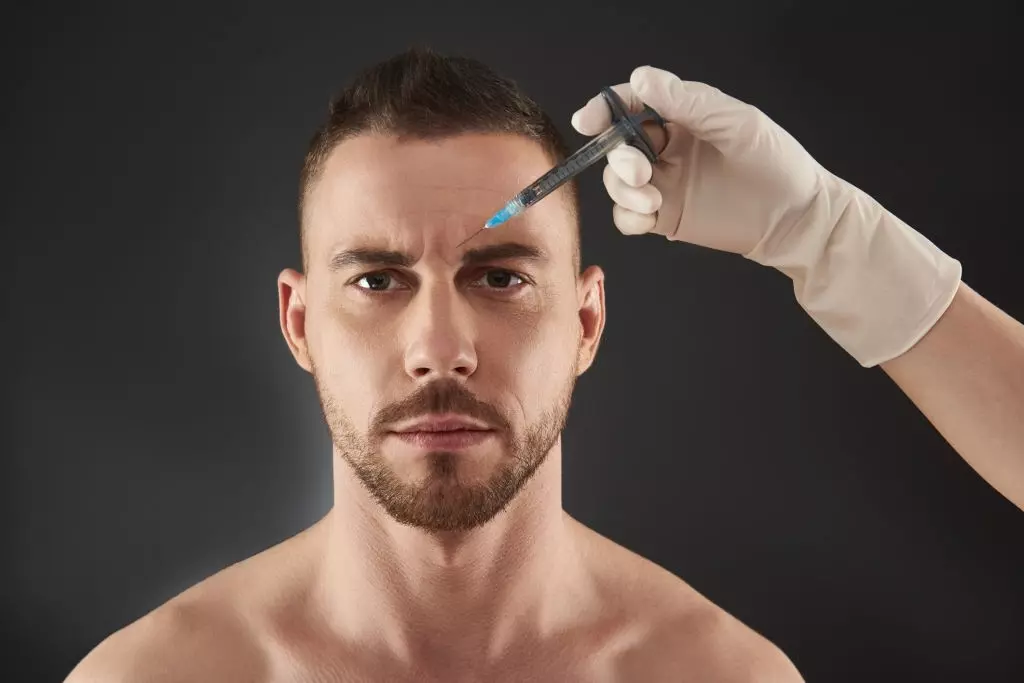 Cintura para cima o retrato de um cara caucasiano olhando para a câmera contra um fundo cinza. Foto recortada do modelo masculino, recebendo injeção de beleza feita por cosmetologista no salão. Conceito de beleza e cuidados com o rosto