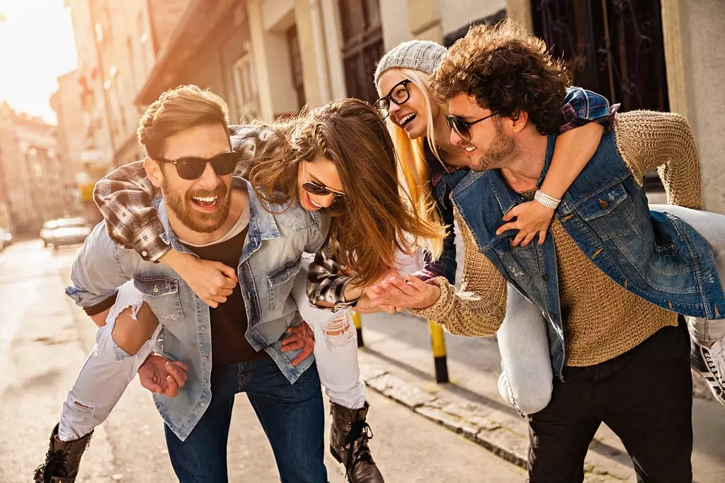 Foto de quatro amigos se divertindo cavalgando nas costas na cidade. Os homens carregam as mulheres e os casais vestem jaqueta jeans, camisa xadrez, boné, óculos e camisa jeans. Eles estão de ótimo humor, rindo e sorrindo, andando em uma pequena rua sem tráfego entre lindas casas antigas.